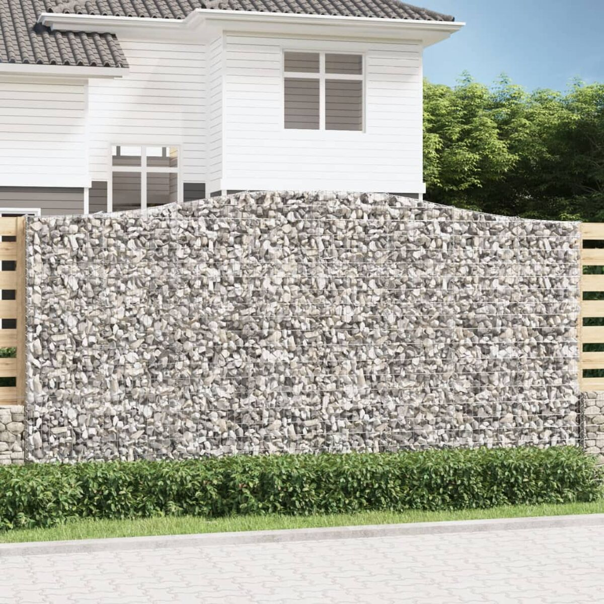 Gabionenmauer mit Steinen, hinter einem Zaun, Haus im Hintergrund.