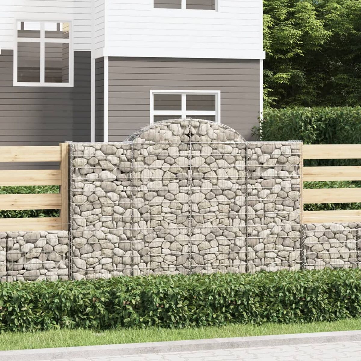 Ein Gabionenwandabschnitt mit einem Holzzaun dahinter und einem Haus im Hintergrund.