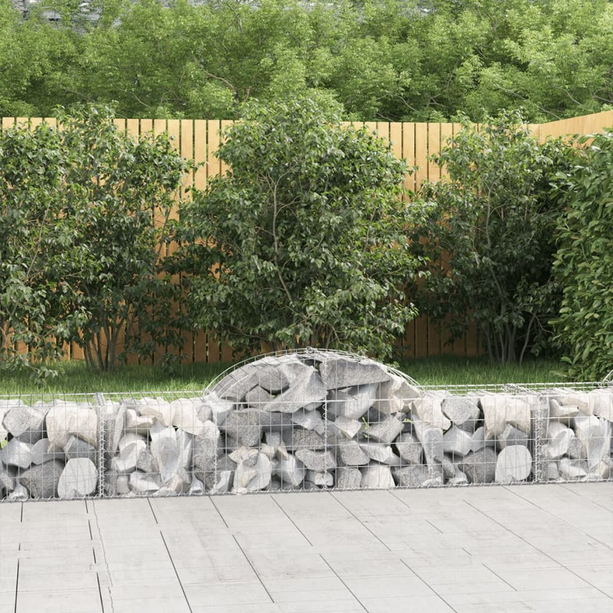 Eine Stein-Gabionenmauer vor grünen Büschen und einem Holzzaun in einem Garten.