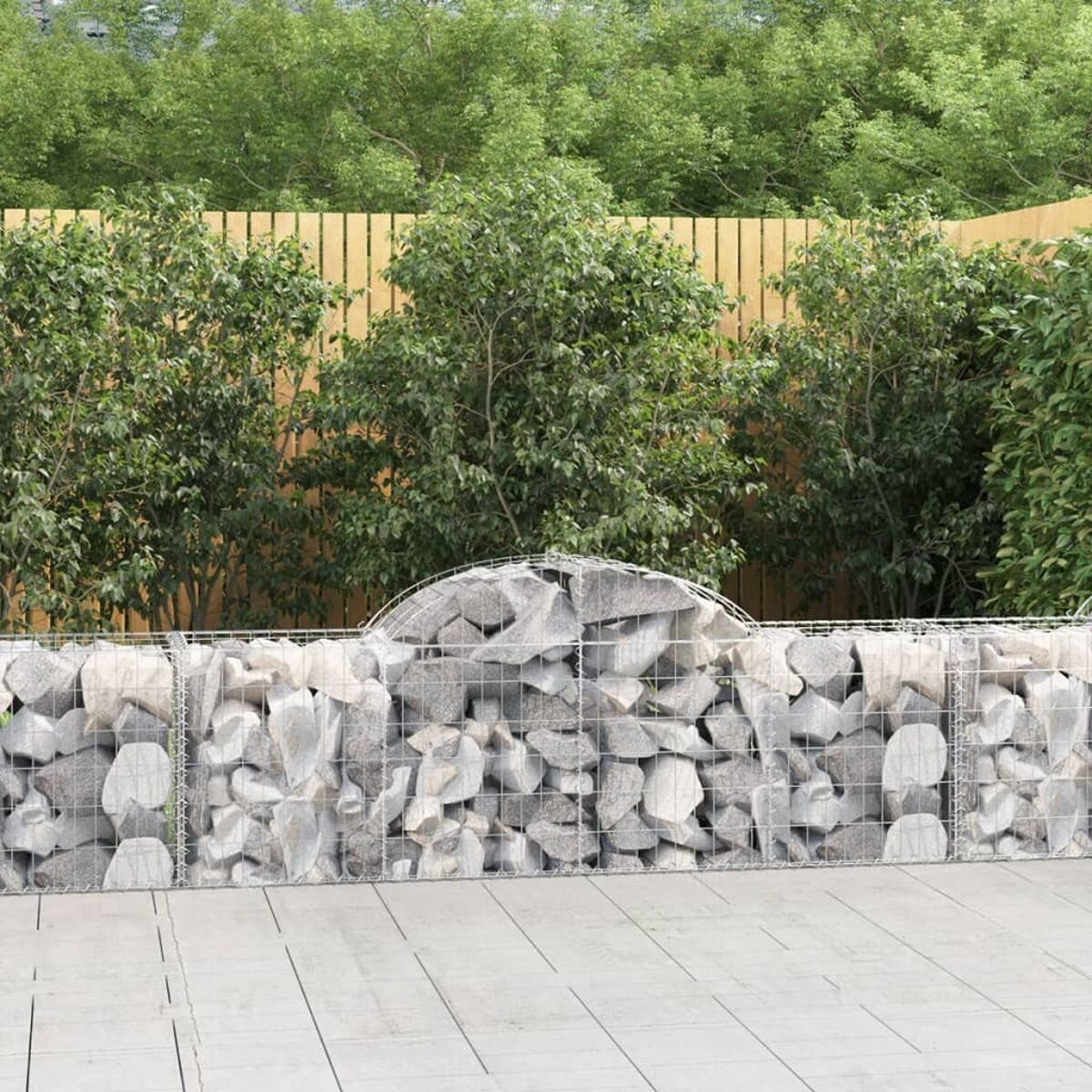 Eine mit Steinen gefüllte Gabionenmauer steht in einem Garten mit Holzzaun und Büschen im Hintergrund.