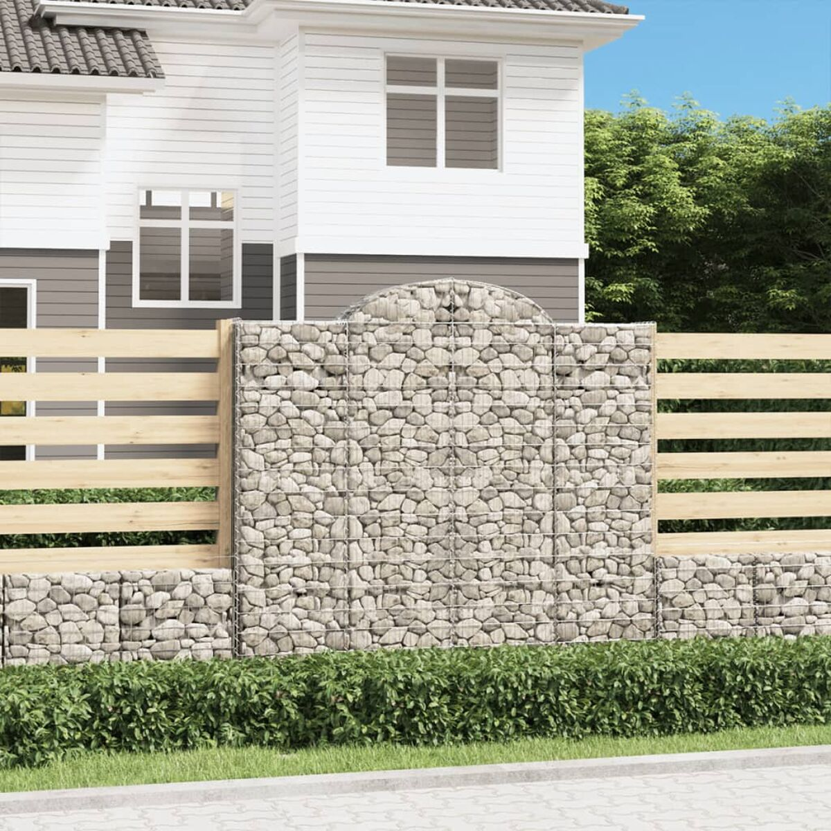 Haus mit Gabionenzaun, steingefüllt, Holzzaun und grünen Büschen davor.