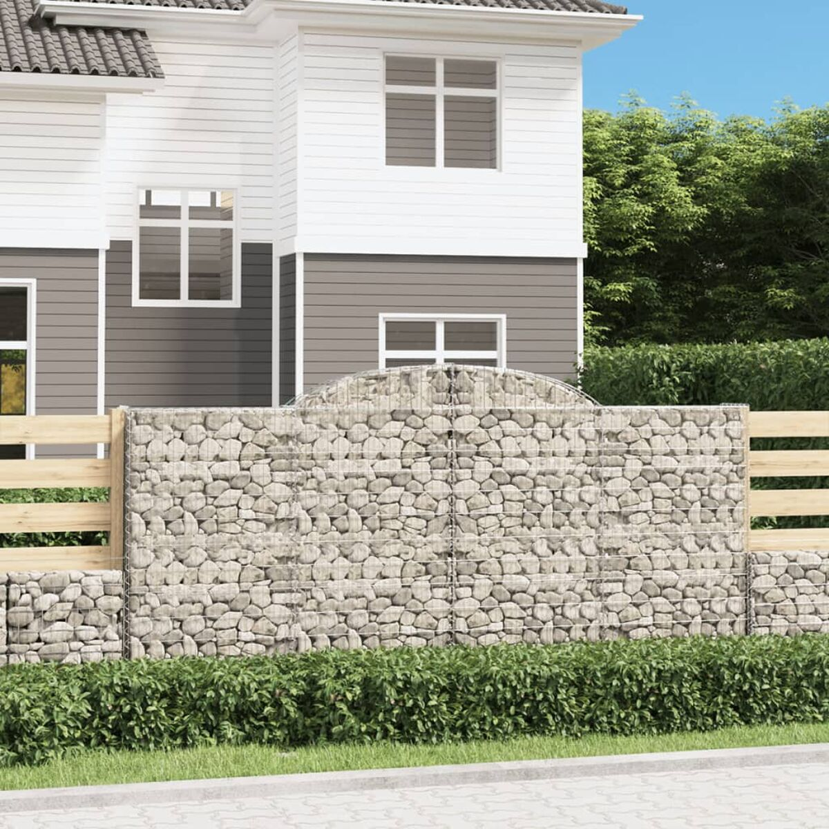 Eine Gabionenwand mit Steinfüllung, Holzzaun und Haus im Hintergrund.