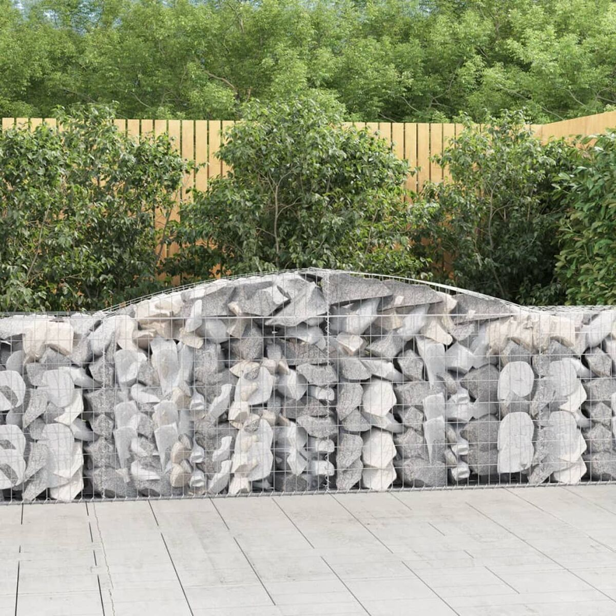 Mit Steinen gefüllte Gabione auf Holzplanken, mit grünen Büschen und Holzzaun im Hintergrund.
