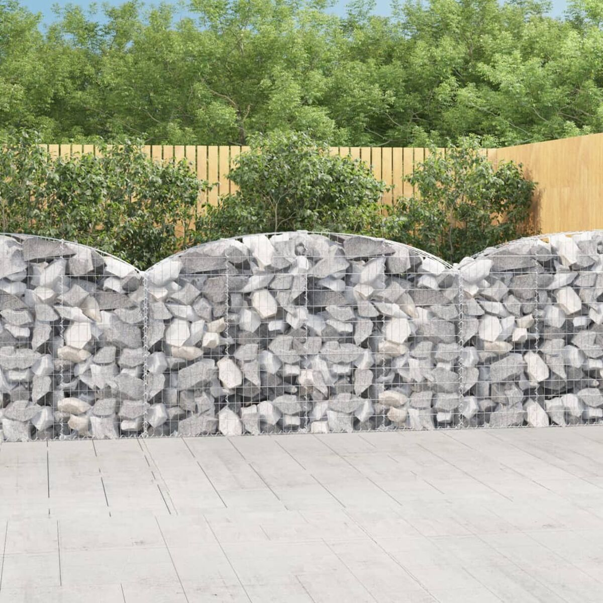 Gabionenmauer, gewölbter Oberteil, Drahtgeflecht gefüllt mit grauen Steinen, in einem Garten. Holzzaun im Hintergrund.