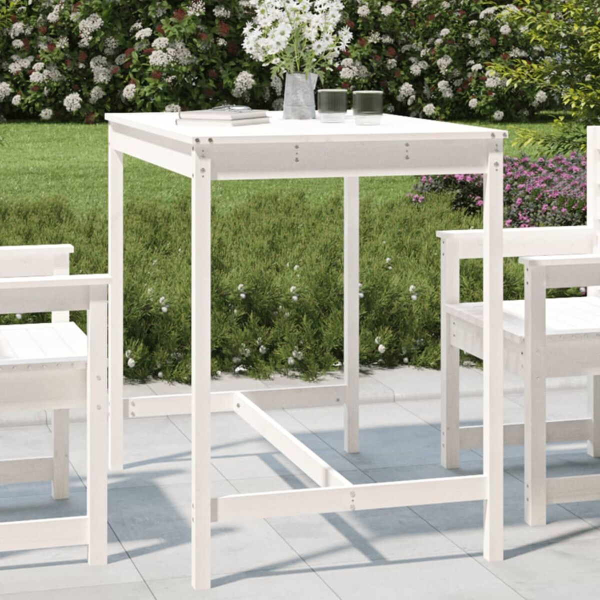 Weißer quadratischer Tisch im Freien mit zwei Stühlen, Blumen oben drauf und Garten im Hintergrund.