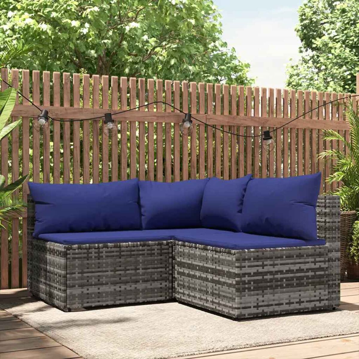 Ein graues, gewebtes Ecksofa mit blauen Kissen. Es steht auf einem Teppich und hat im Hintergrund einen Holzzaun und Lichter.