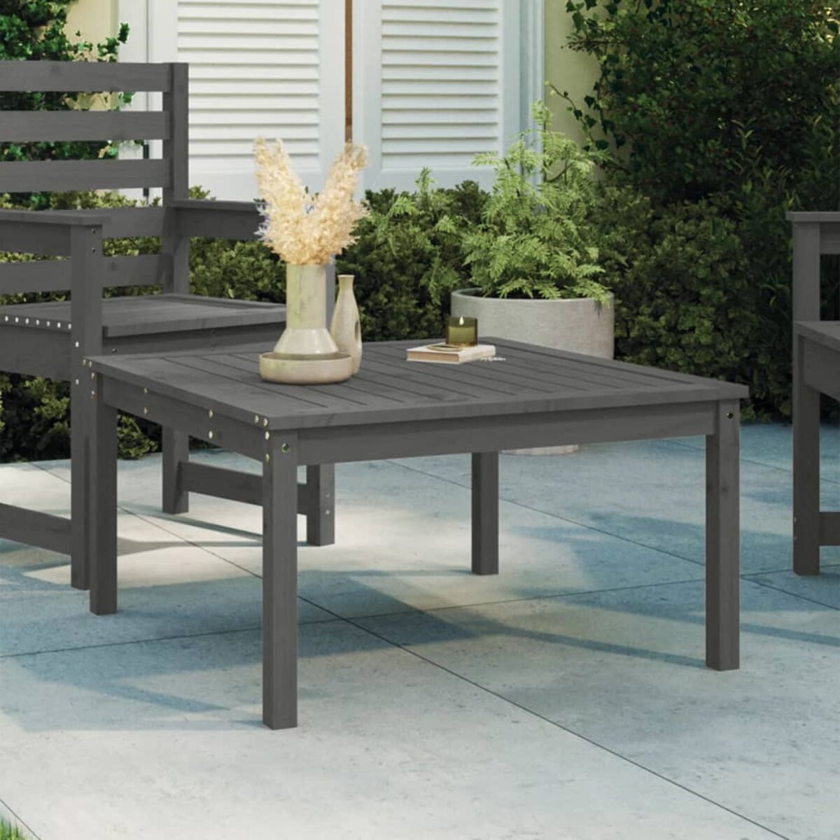Grauer Holzkaffeetisch im Freien, Stuhl und Vase mit Pflanzen. Beige Terrasse mit Grün im Hintergrund.