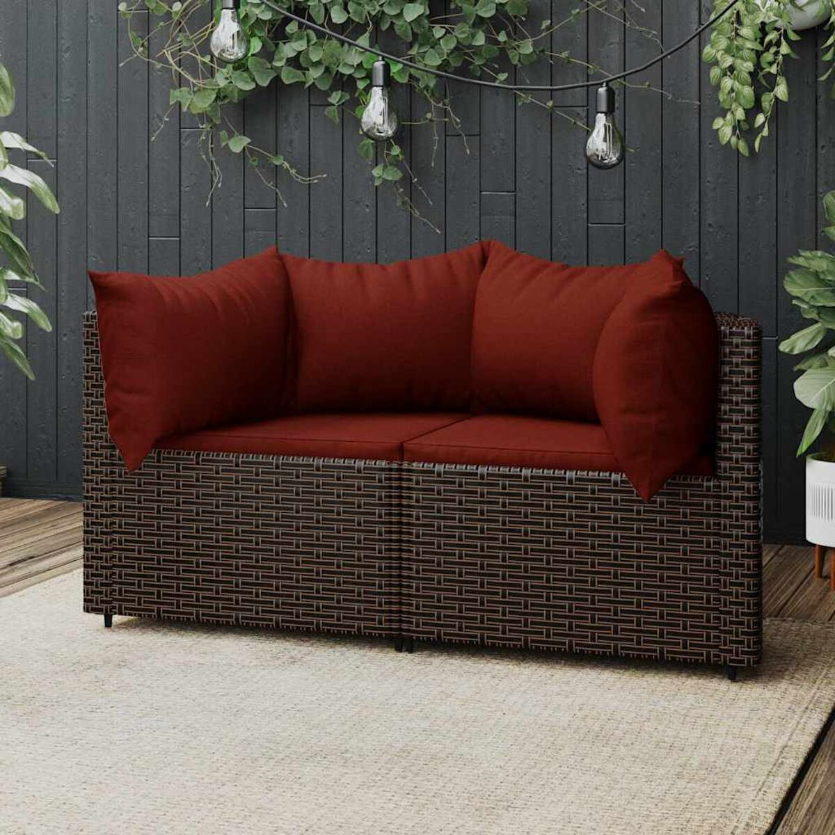 Ein zweisitziges braunes Korbmöbelsofa mit roten Kissen auf einem Teppich mit Pflanzen.