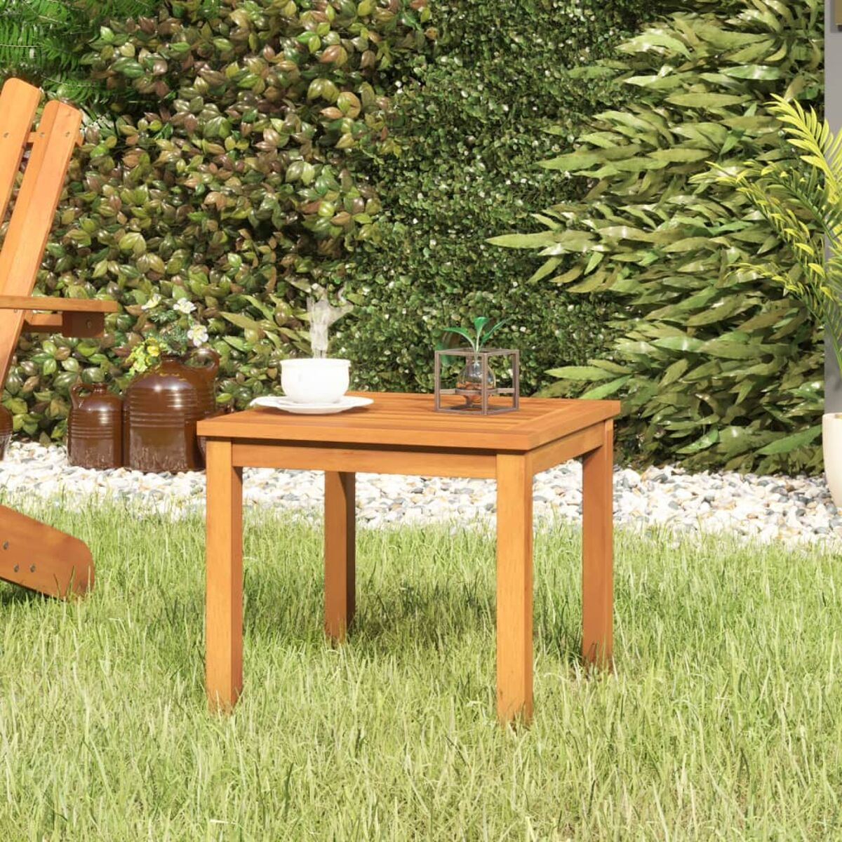 Quadratischer Holztisch mit Tasse. Gartenhintergrund mit grünem Gras und Büschen. Ein Stuhl ist im Hintergrund sichtbar.
