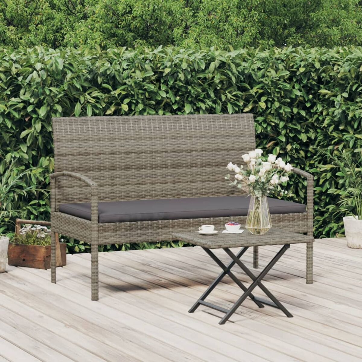 Weidenbank, Tisch mit Blumen und zwei Tassen auf Holzterrasse, grüner Heckenhintergrund.