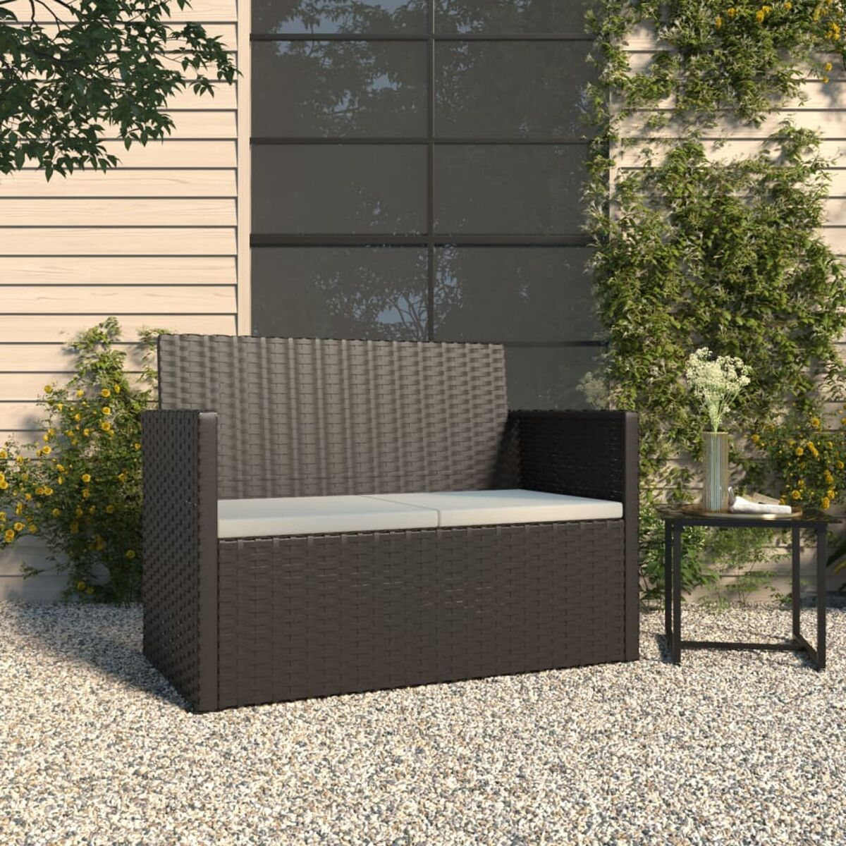 Eine braune, gewebte Outdoor-Bank mit einem kleinen Tisch, auf Kies, an einer Wand.