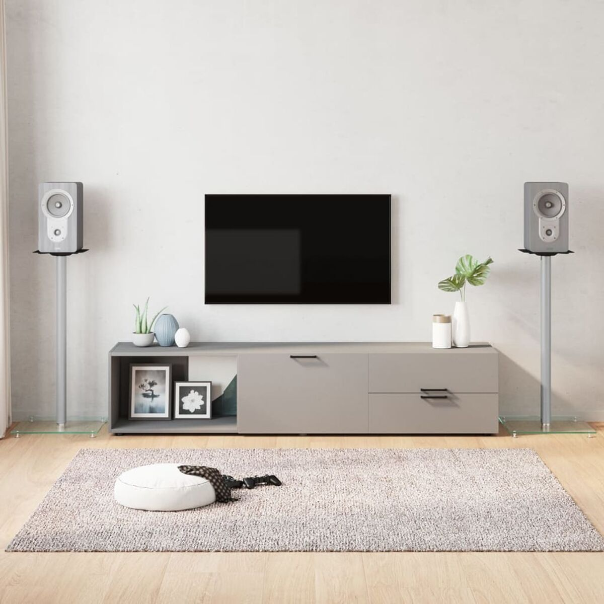 Wohnzimmer-Setup: Fernseher auf einem grauen Schrank, Lautsprecher auf Ständern, Teppich und Pouf.