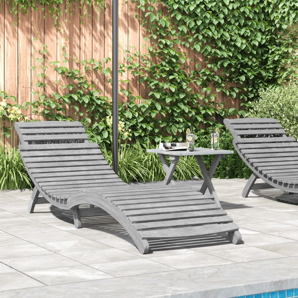 Due lettini di legno su un patio con un tavolino e fogliame, giornata di sole.