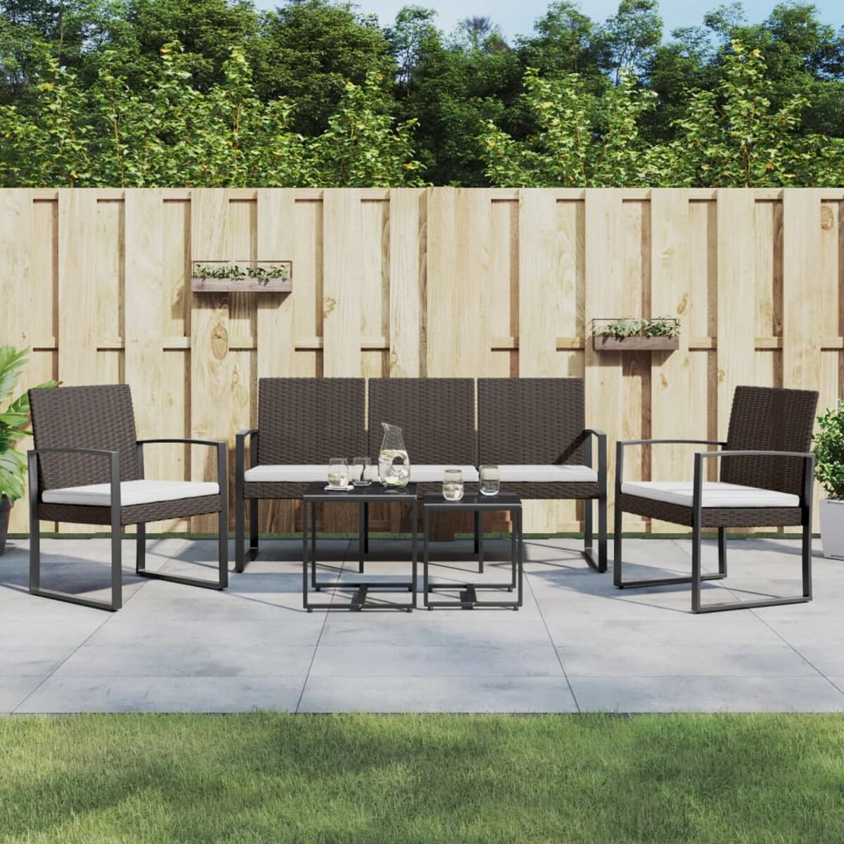 Outdoor-Sitzgruppe. Dunkelbraunes Sofa und zwei Stühle, kleine Tische. Holzzaun und grüne Pflanzen.