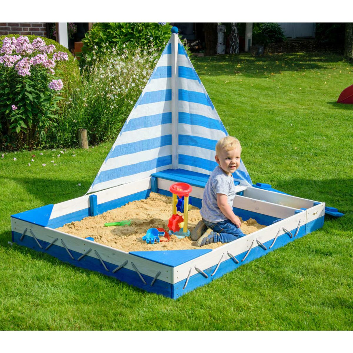 Ein Kind in einem Sandkasten mit einem blau-weiß gestreiften Segel. Grünes Gras.