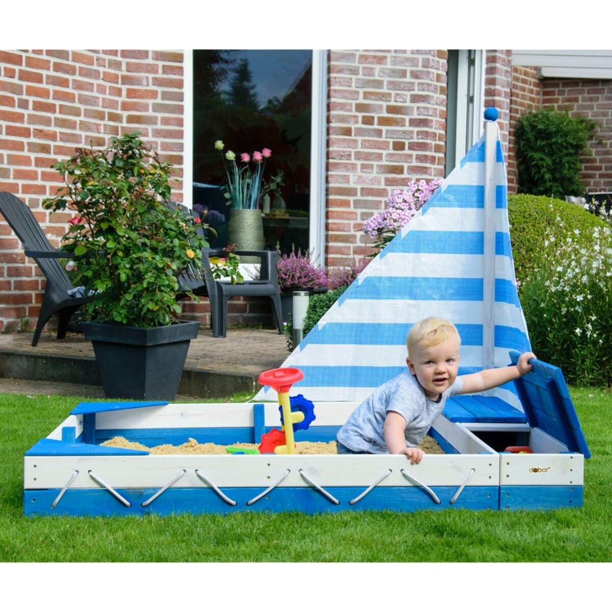 Kind in einem blauen Sandkasten in Bootsform. Rotes Spielzeug liegt im Sand.