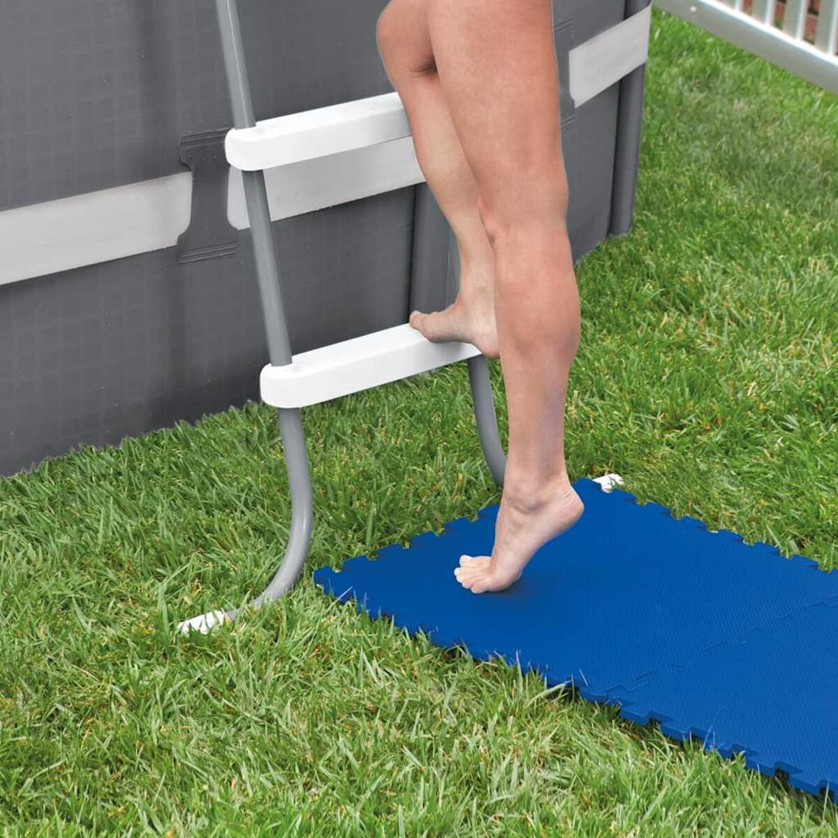 Person betritt einen Pool. Beine sind sichtbar, mit einer Poolleiter. Blaue Matte auf Gras. Graue Poolseite.