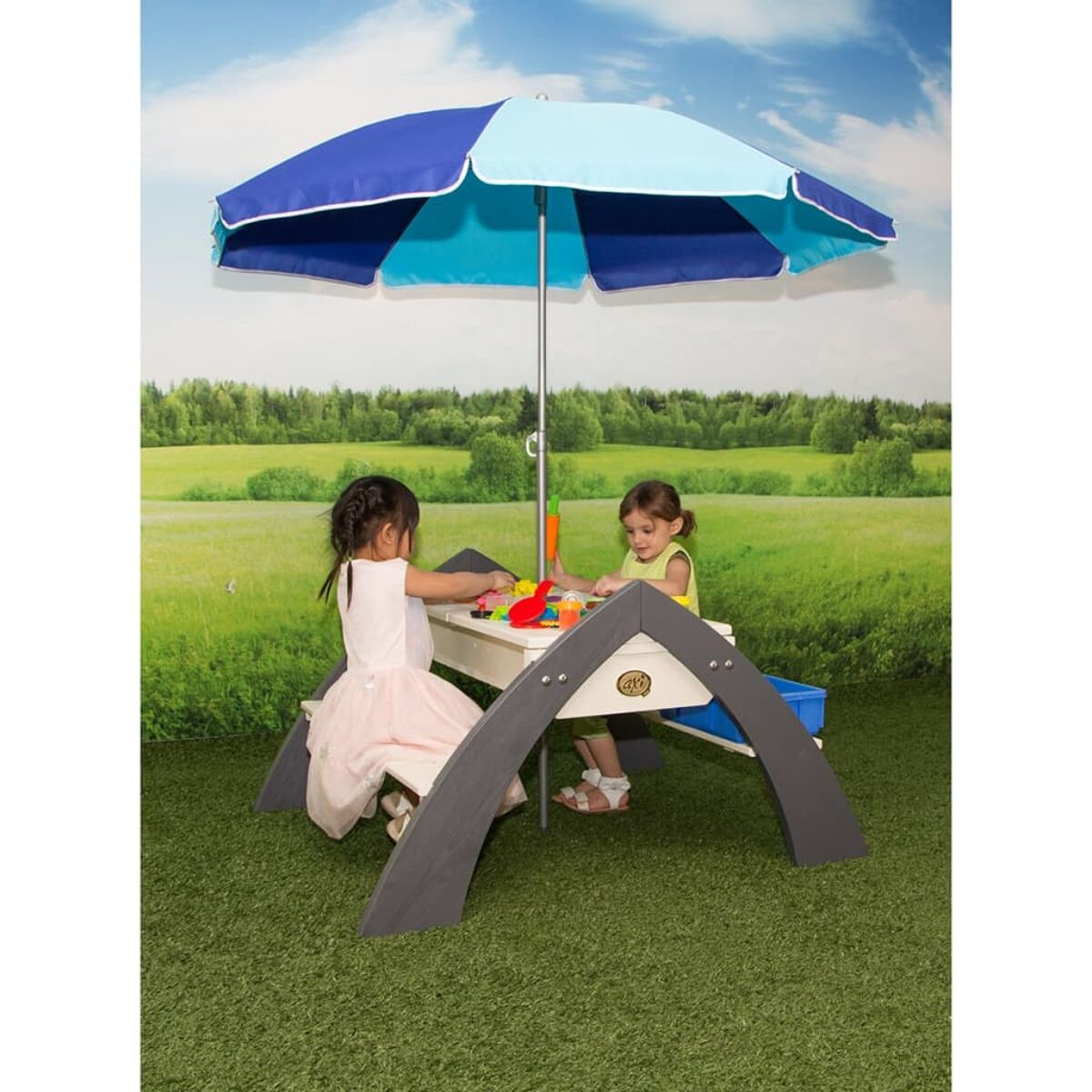 Zwei Mädchen spielen an einem weißen Picknicktisch unter einem blau-hellblauen Sonnenschirm auf grünem Gras.