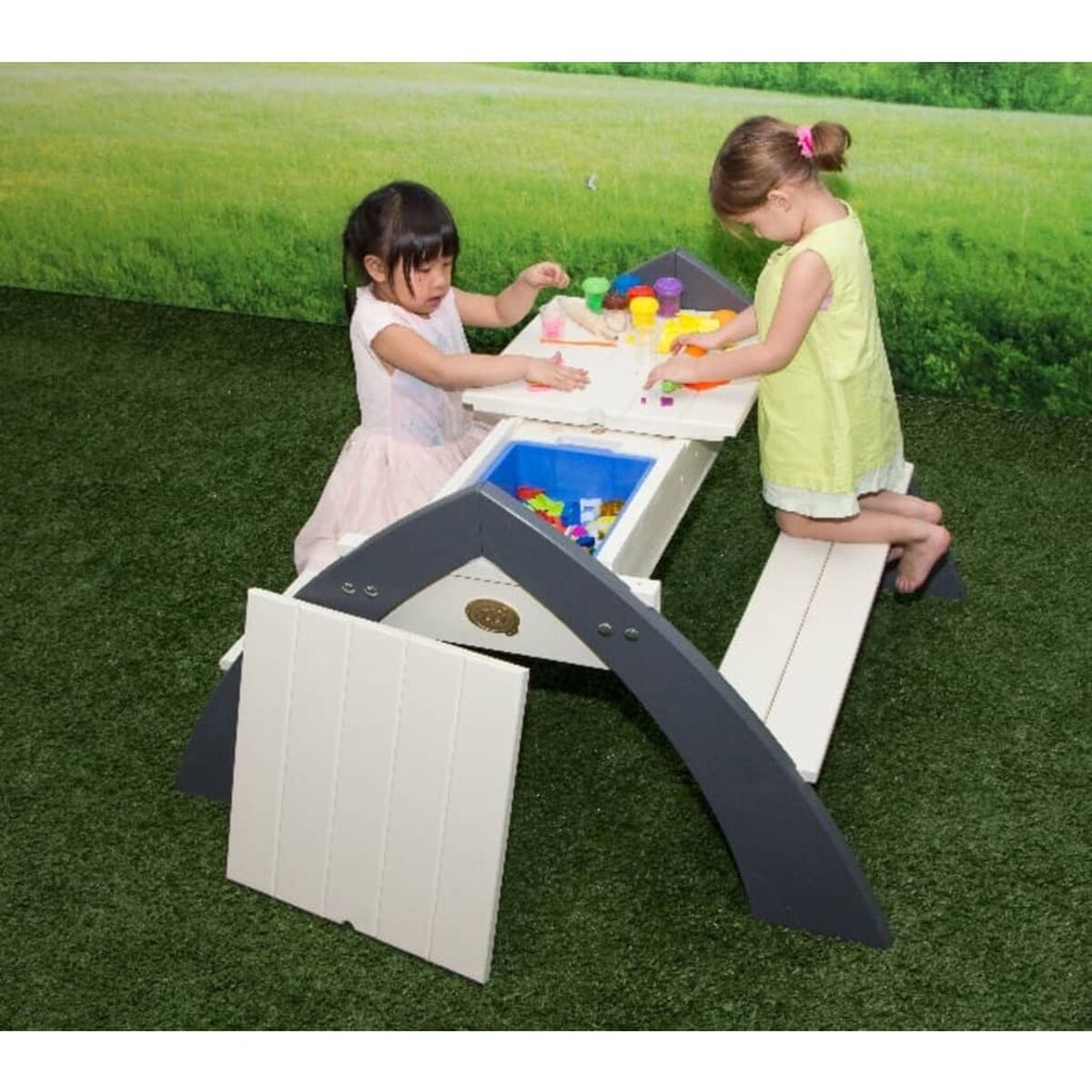 Zwei Mädchen spielen draußen an einem Kinderspieltisch. Grüner Rasen im Hintergrund. Weißer und grauer Tisch.