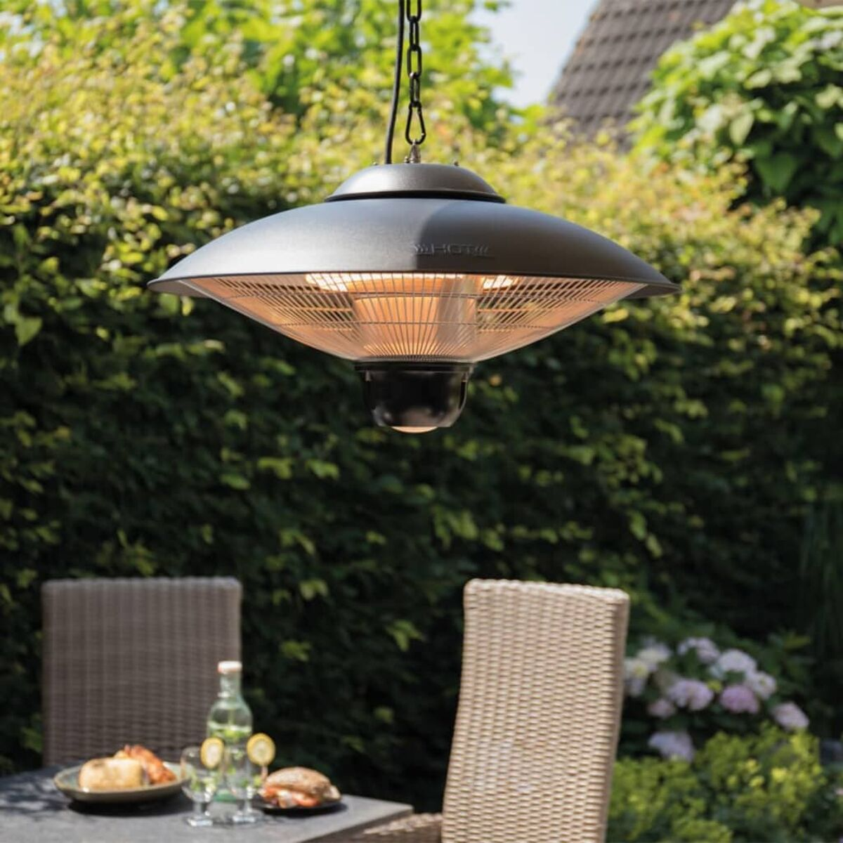 Outdoor scene with a hanging black heater, table with food and drinks, and green foliage in background.