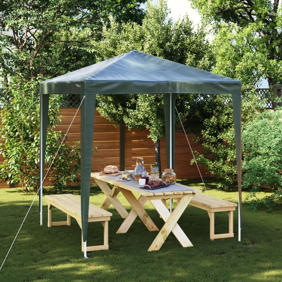 Pavillon mit Tisch und Bänken in einem grünen Garten, Bäume im Hintergrund.