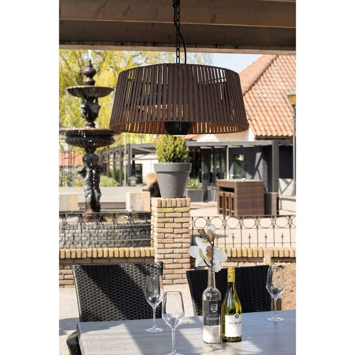 Ein brauner Lampenschirm hängt über einem Tisch mit Gläsern und Flaschen. Ein Brunnen ist im Hintergrund.