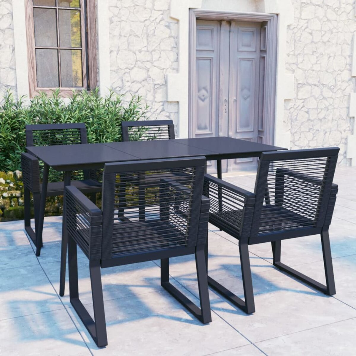 Schwarze Essgruppe im Freien auf einer Terrasse. Enthält einen Tisch und Stühle mit gewebten Details. Hintergrund zeigt Steinhaus.