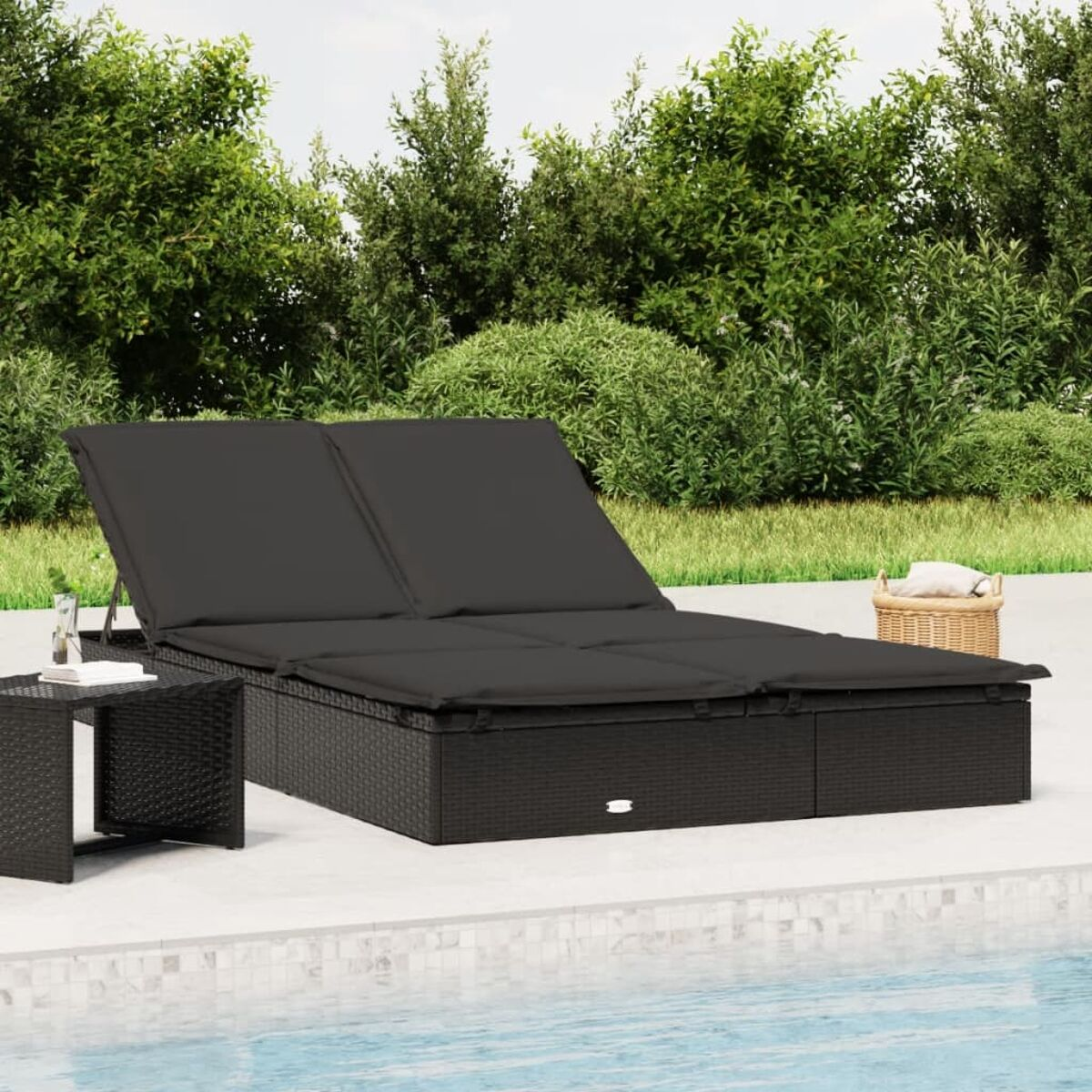 Schwarze Outdoor-Doppelliege mit Kissen. Kleiner Tisch und Korb. Grüne Büsche im Hintergrund.