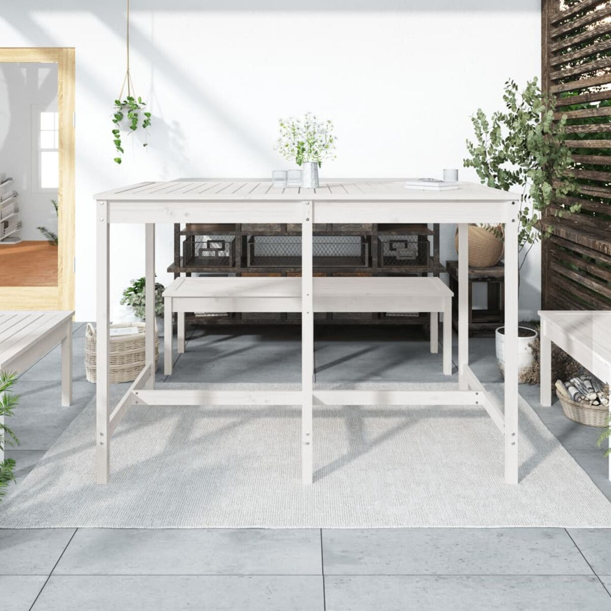 Weißes hölzernes Outdoortisch- und Bankset auf einer Terrasse mit Pflanzen. Der Hintergrund zeigt eine weiße Wand und Tür.