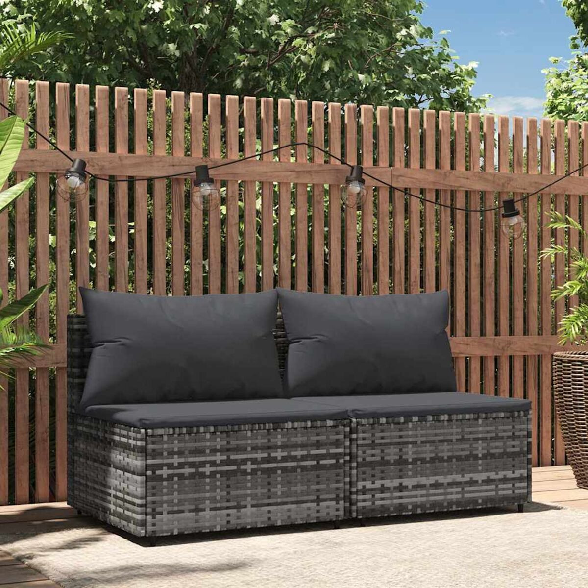 Graues Outdoor-Sofa aus Korbgeflecht mit zwei Kissen. Holzzaun und Lichterkette.