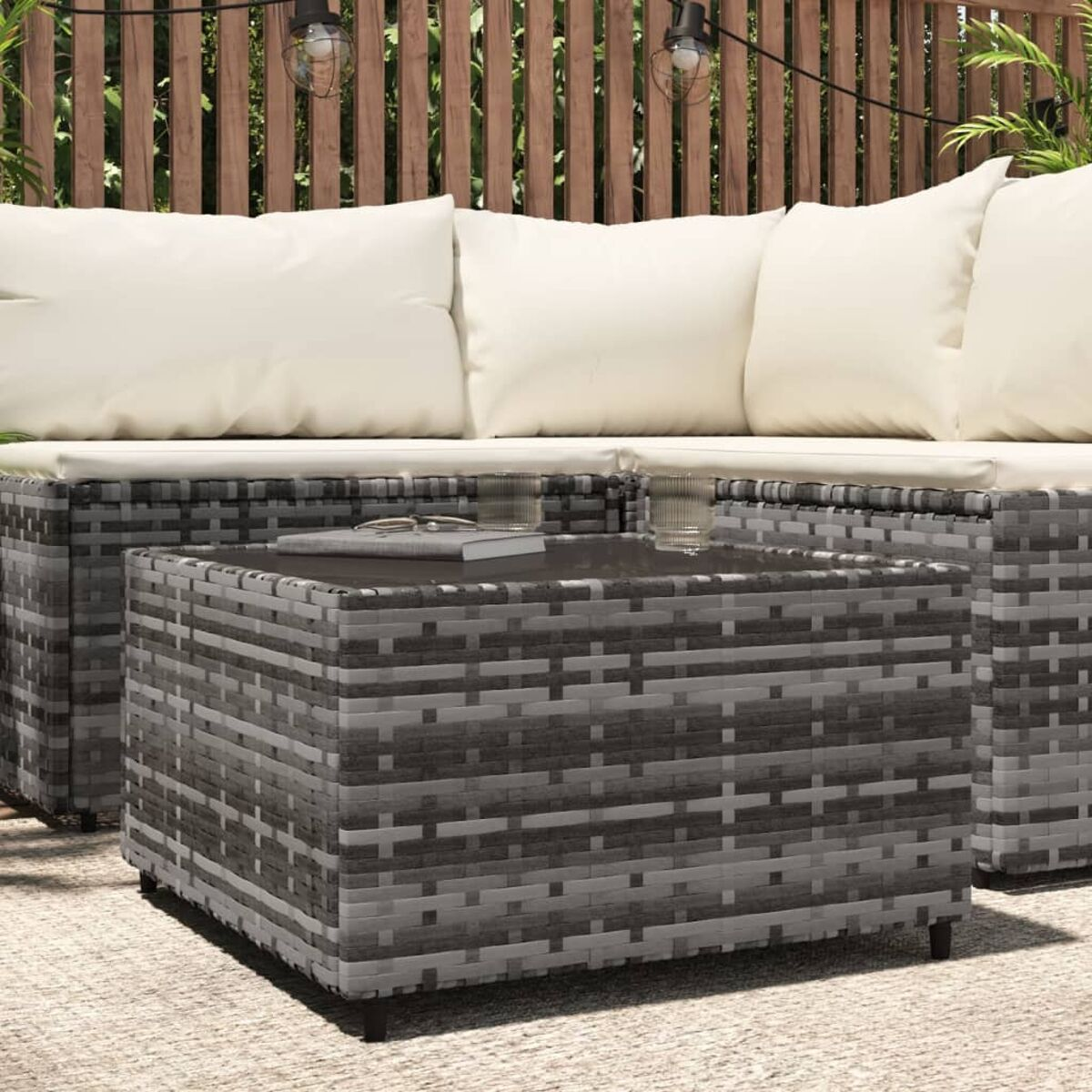 Grauer Korbtisch mit Glasplatte. Er steht auf einer Terrasse mit Sofa und Pflanzen.