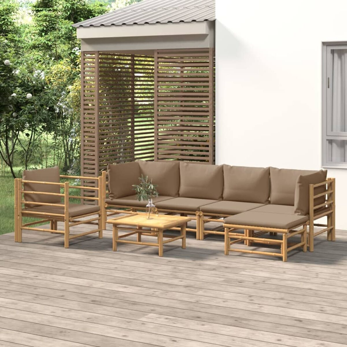 Ein Bambus-Gartenmöbel-Set: Sofa, Stuhl und Tisch. Das Set steht auf einer Holzterrasse. Hintergrund: ein Haus.