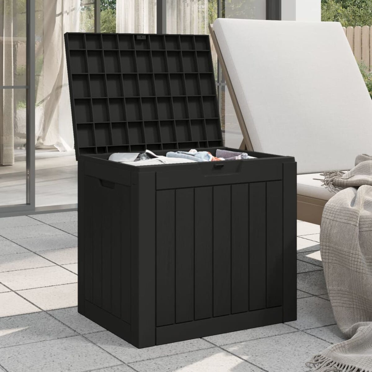 Schwarze Aufbewahrungsbox mit offenem Deckel, auf gefliester Oberfläche mit weißer Sonnenliege im Hintergrund.