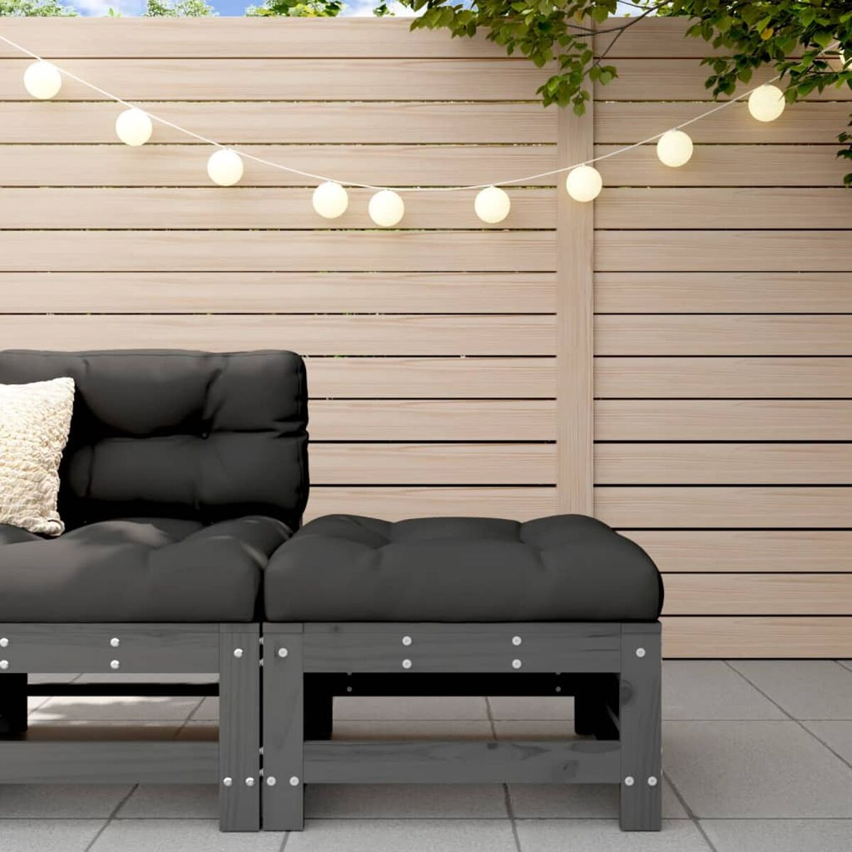 Ein dunkelgraues Sofa im Freien mit weißen Kissen. Ein Holzzaun und Lichterketten sind im Hintergrund.