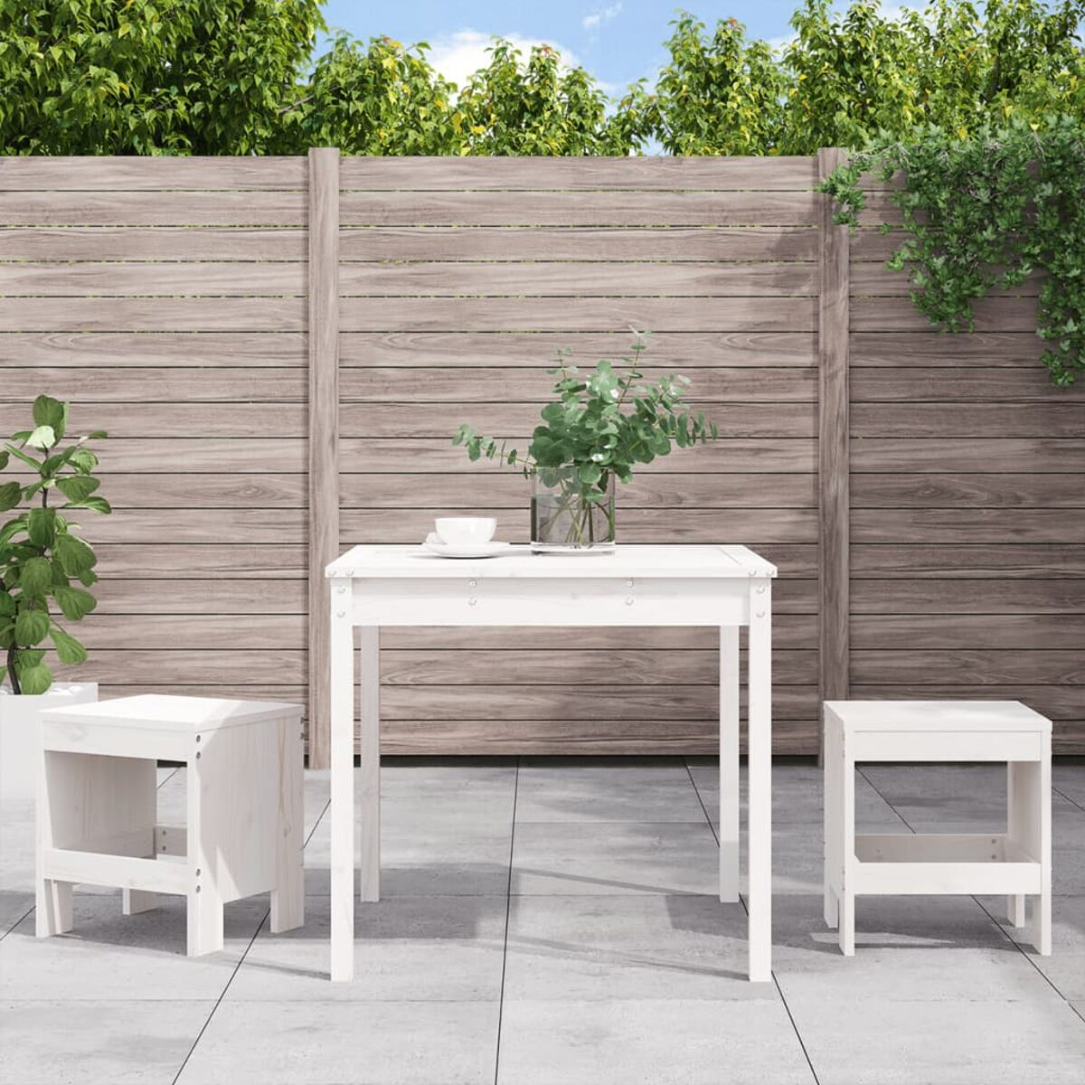 Weißer Gartentisch und zwei Hocker. Grüne Pflanzen und Holzzaun im Hintergrund, vor blauem Himmel.