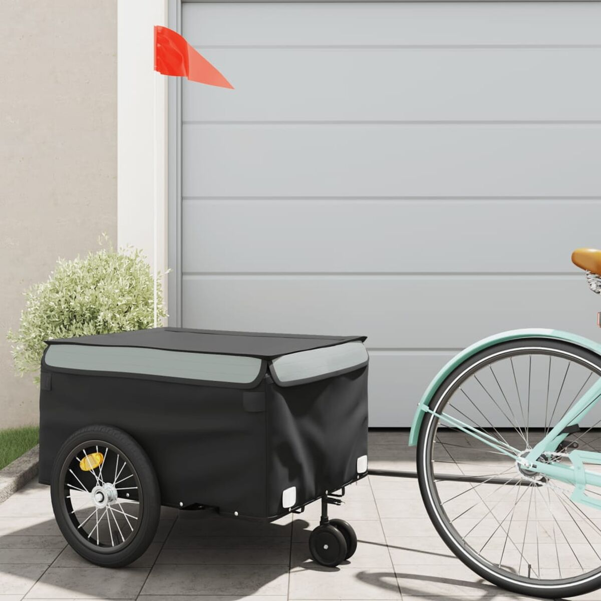 Fahrradanhänger mit schwarzer Box, roter Flagge. Ein Fahrrad im Hintergrund.