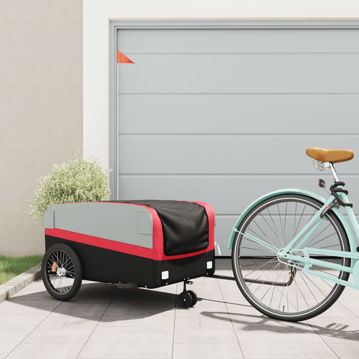 Fahrrad mit Anhänger. Anhänger ist schwarz, rot und grau. Geparkt auf Beton neben einer Garage.