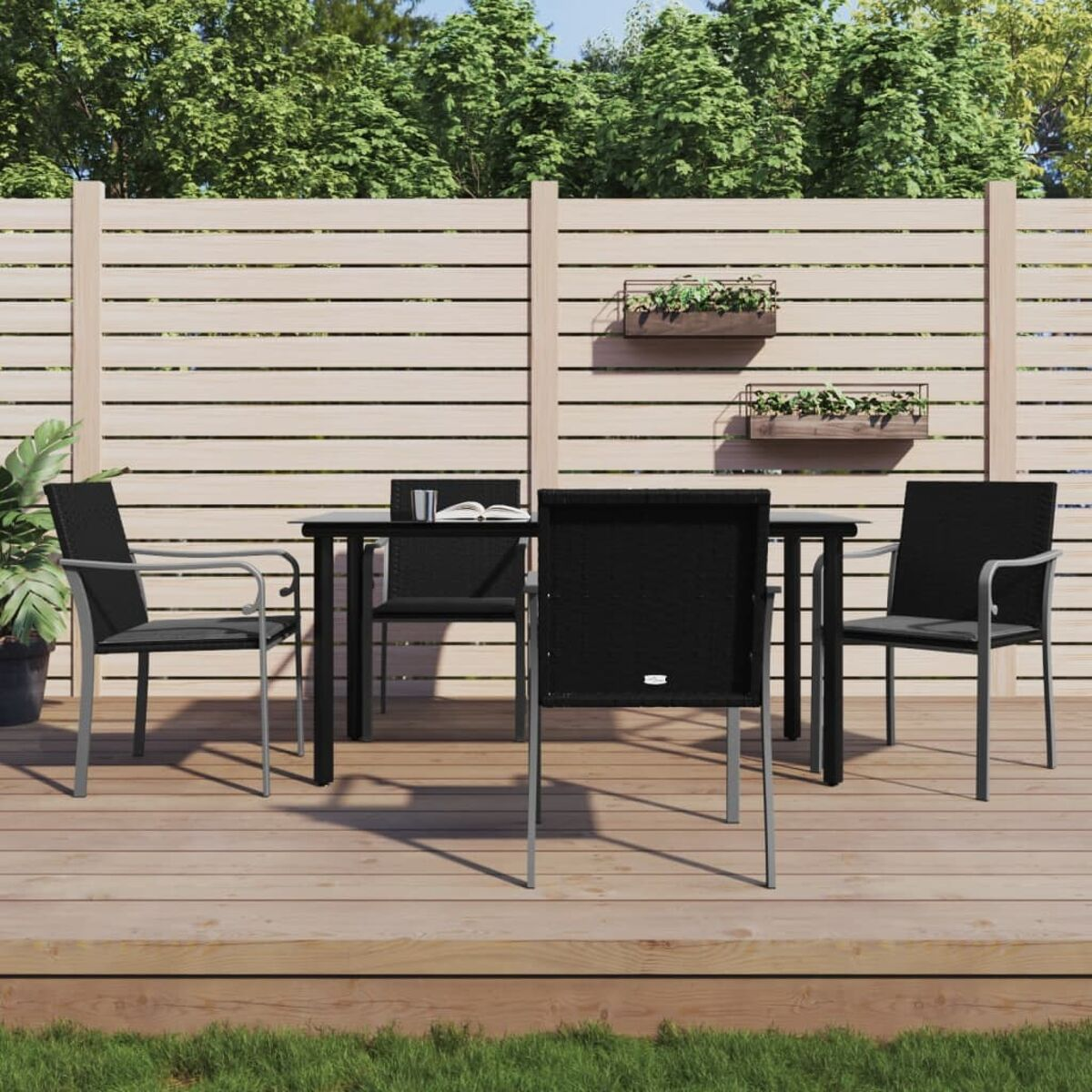 Essgruppe im Freien mit schwarzem Tisch und Stühlen auf Holzterrasse. Grüne Pflanzen und Holzzaun im Hintergrund.