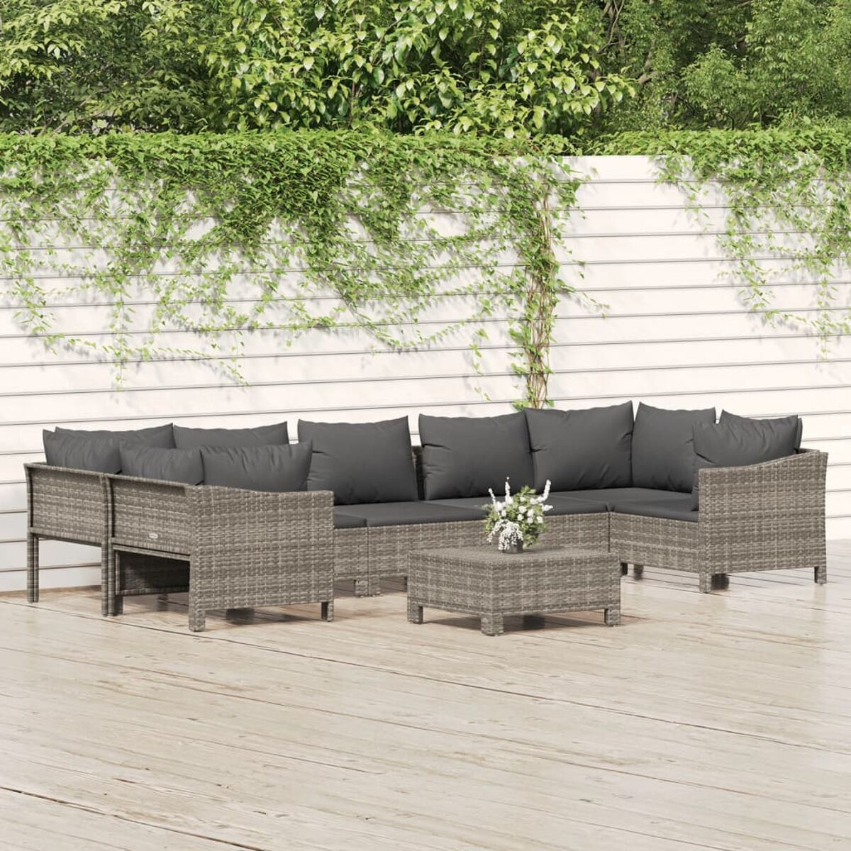 Graues Rattan-Gartenmöbelset mit grauen Kissen, Couchtisch mit Blumen, Holzterrasse und Laub.