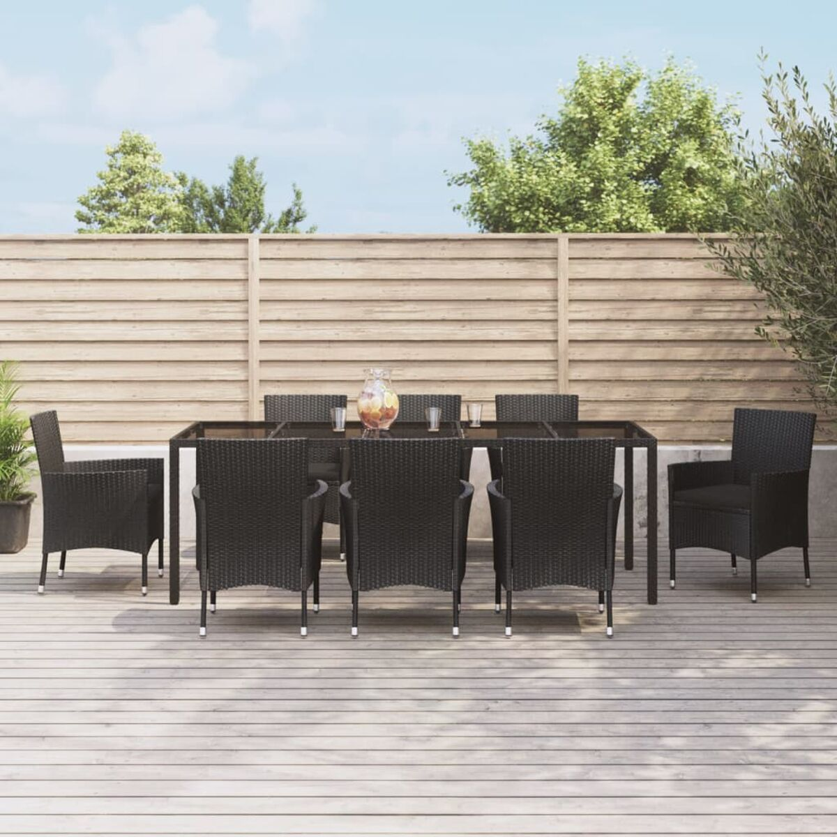Gartenmöbel mit Tisch, acht Stühlen und einem Krug. Holzterrasse, Zaun und Bäume im Hintergrund.