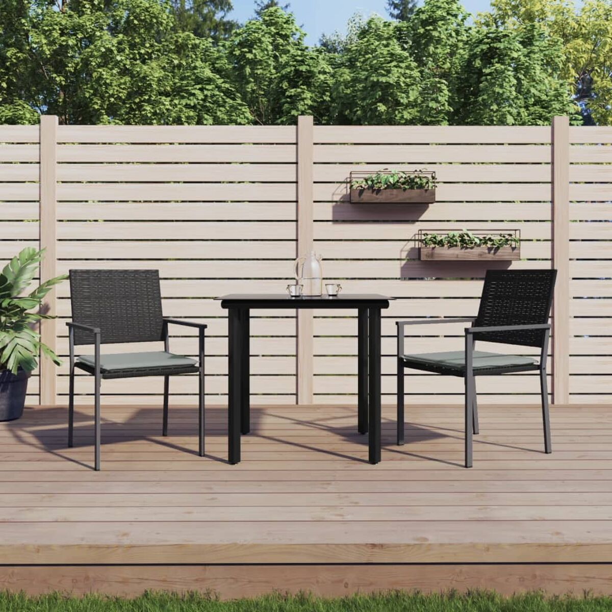 Ein schwarzer Tisch und zwei Stühle auf einer Holzterrasse, Pflanzen im Hintergrund.