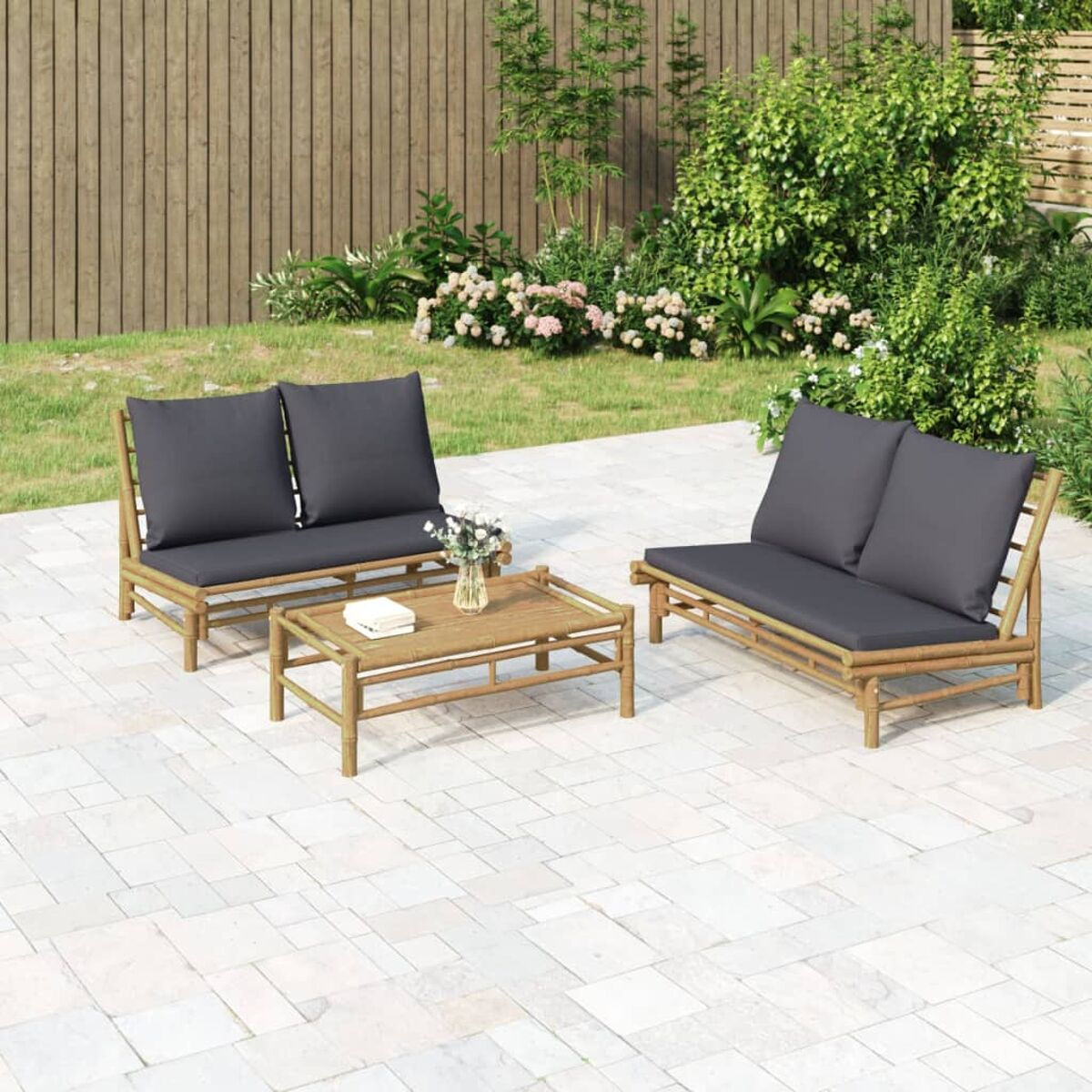 Outdoor-Bambusmöbel-Set mit grauen Kissen auf gepflasterter Fläche, mit Pflanzen und Zaun.
