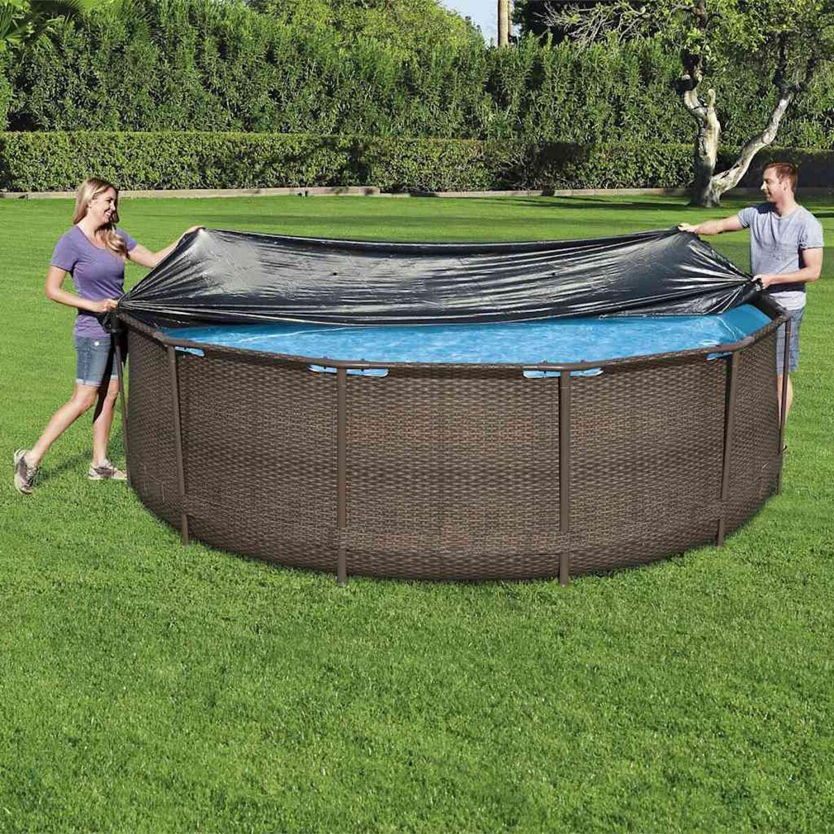 Zwei Personen bedecken einen runden, braunen Pool mit einer schwarzen Abdeckung, auf grünem Gras, umgeben von Bäumen.