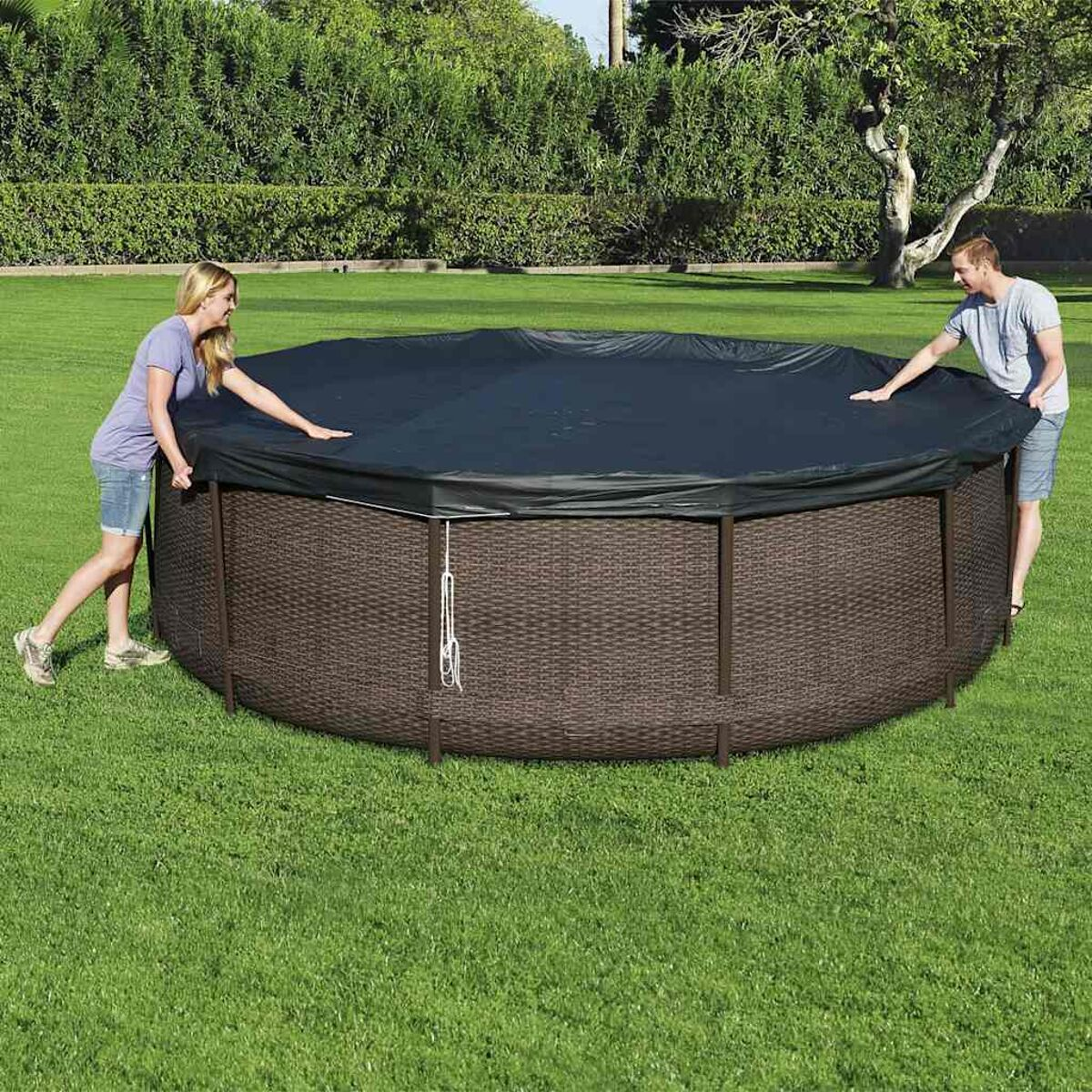 Zwei Personen decken einen runden, braunen Pool mit dunkler Abdeckung auf einer grünen Wiese ab.
