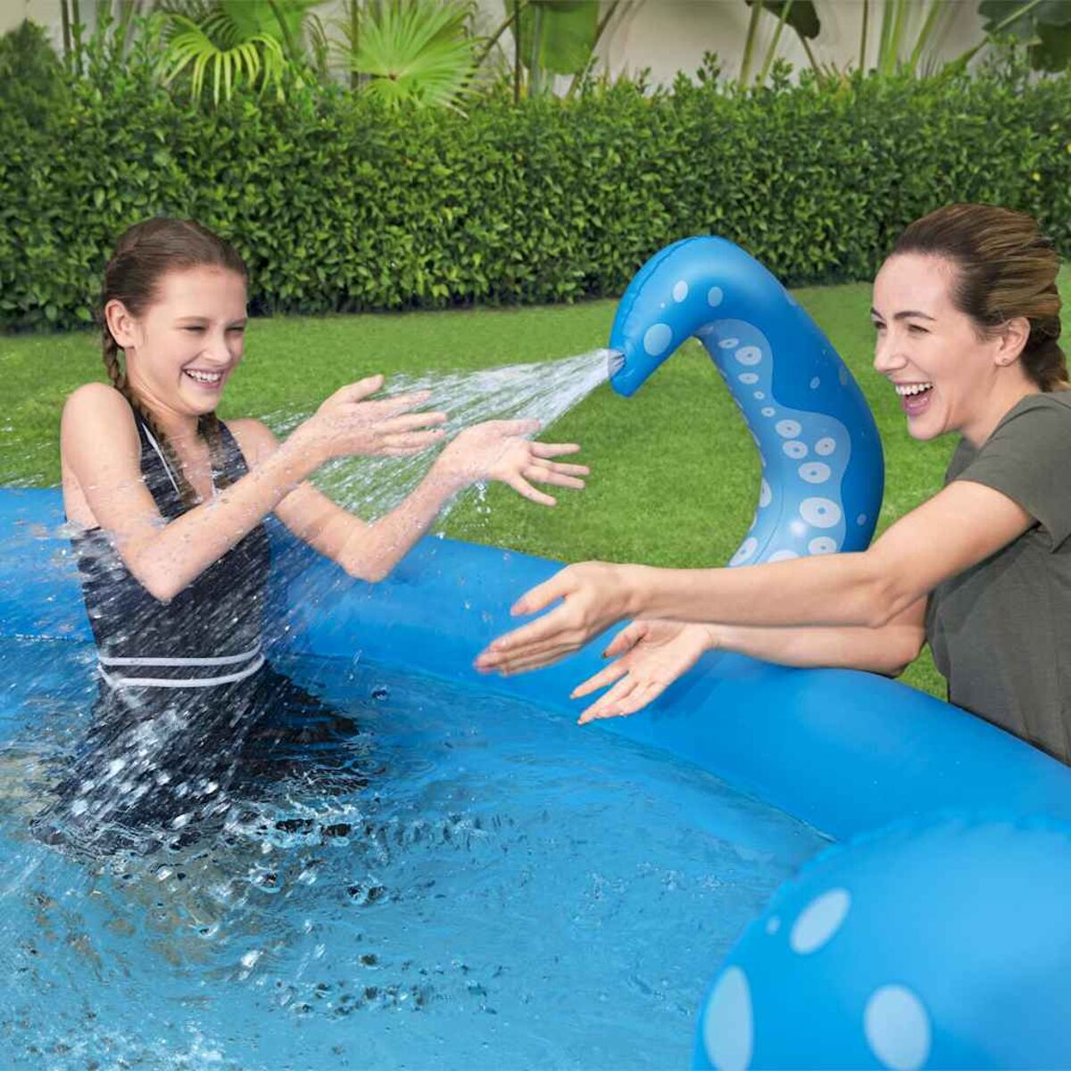 Zwei Frauen spielen in einem blauen Pool mit einem Wasserspiel, das Wasser versprüht.