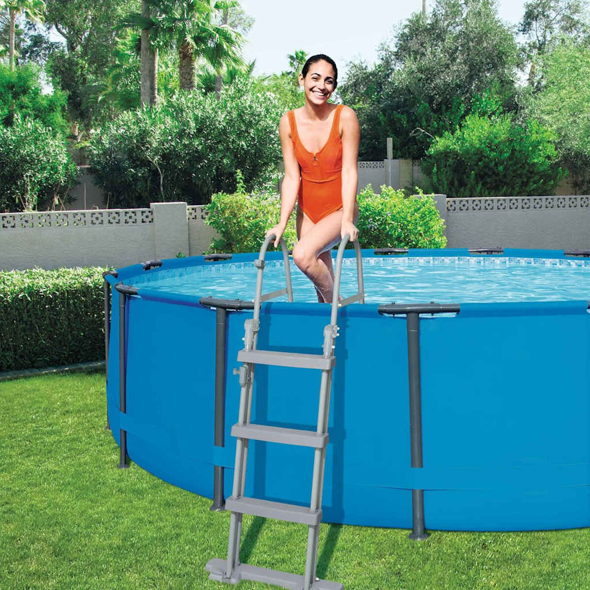 Frau verlässt einen blauen Aufstellpool über eine Leiter, umgeben von grünem Gras und Bäumen.