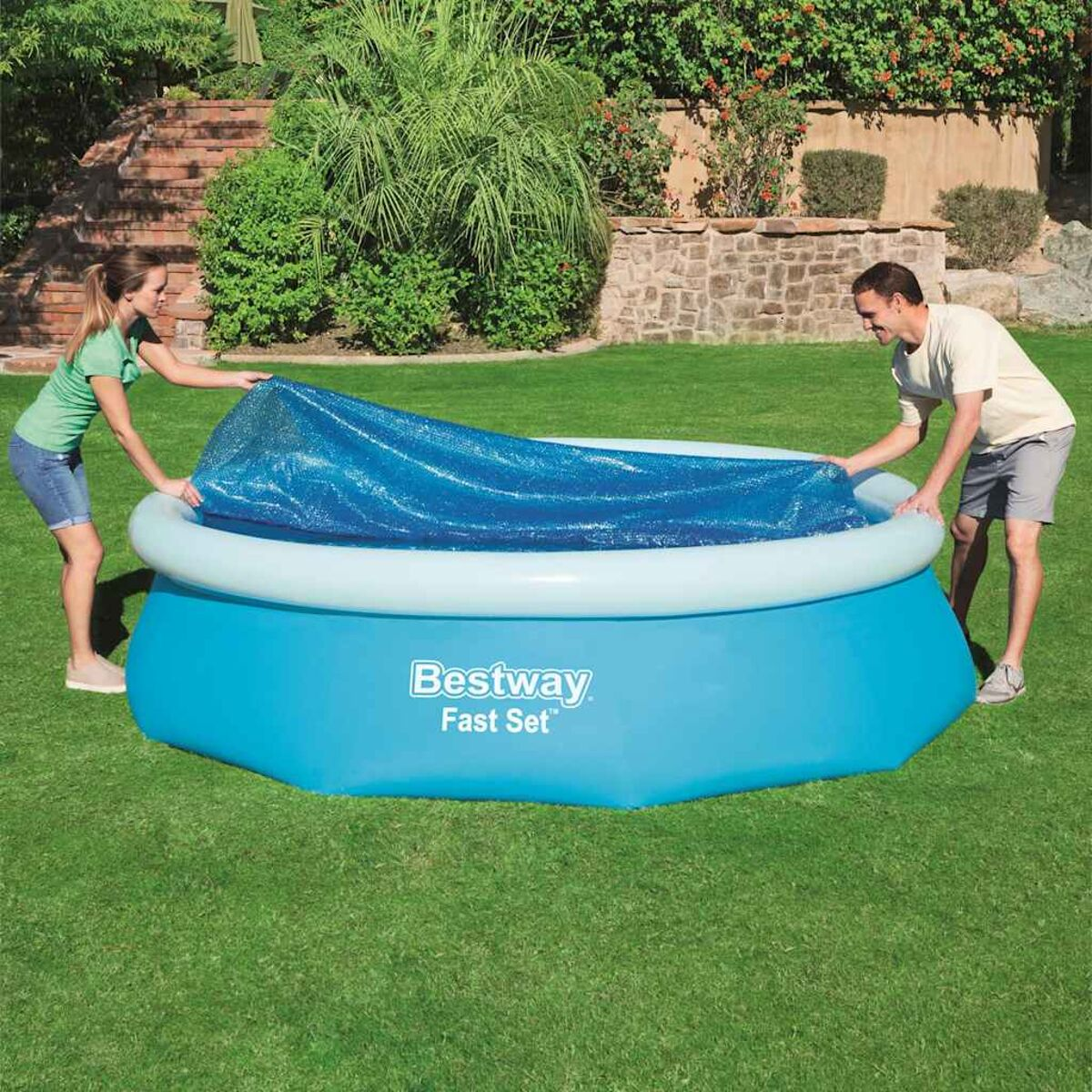 Zwei Personen bedecken einen blauen Pool mit einer blauen Abdeckung in einem grünen Garten.