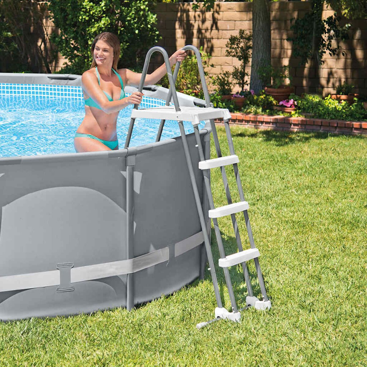 Eine Frau in einem Bikini steht in einem Pool und benutzt eine Leiter. Der Pool befindet sich in einem grasbewachsenen Garten.