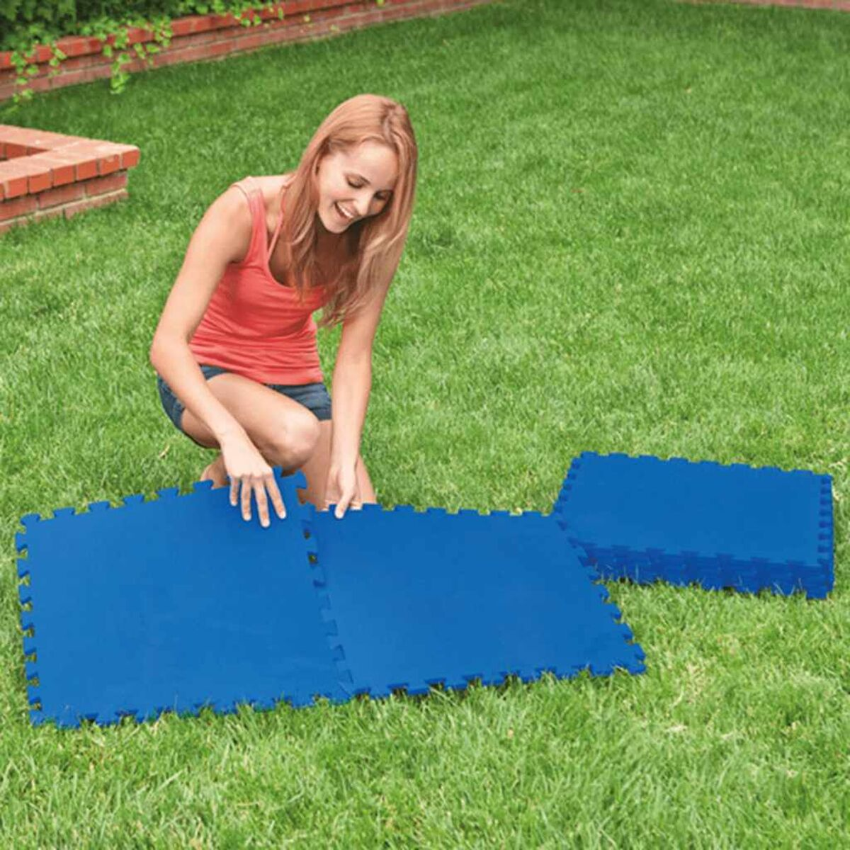 Frau kniet und arrangiert blaue, ineinandergreifende Matten auf grünem Gras, sonniger Tag.