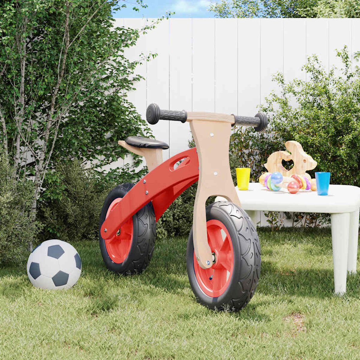 Rotes und hölzernes Laufrad auf Gras. Weißer Tisch mit Spielzeug und Getränken im Hintergrund. Grünes Gras.